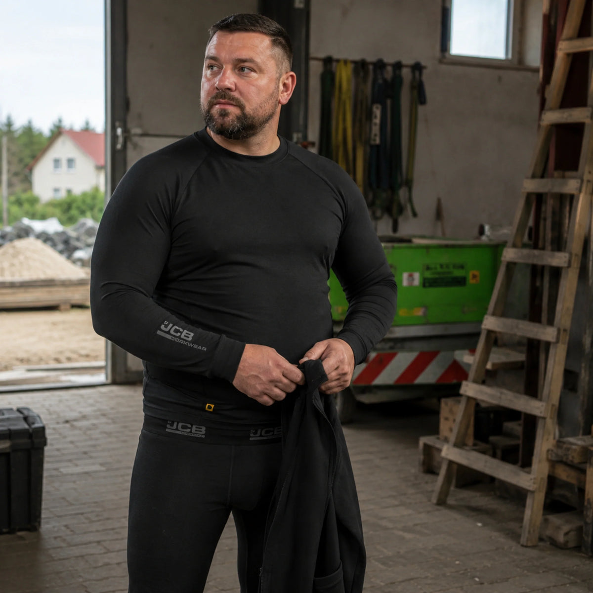 Mężczyzna w czarnej koszulce termoaktywnej JCB w warsztacie, ujęcie z przodu