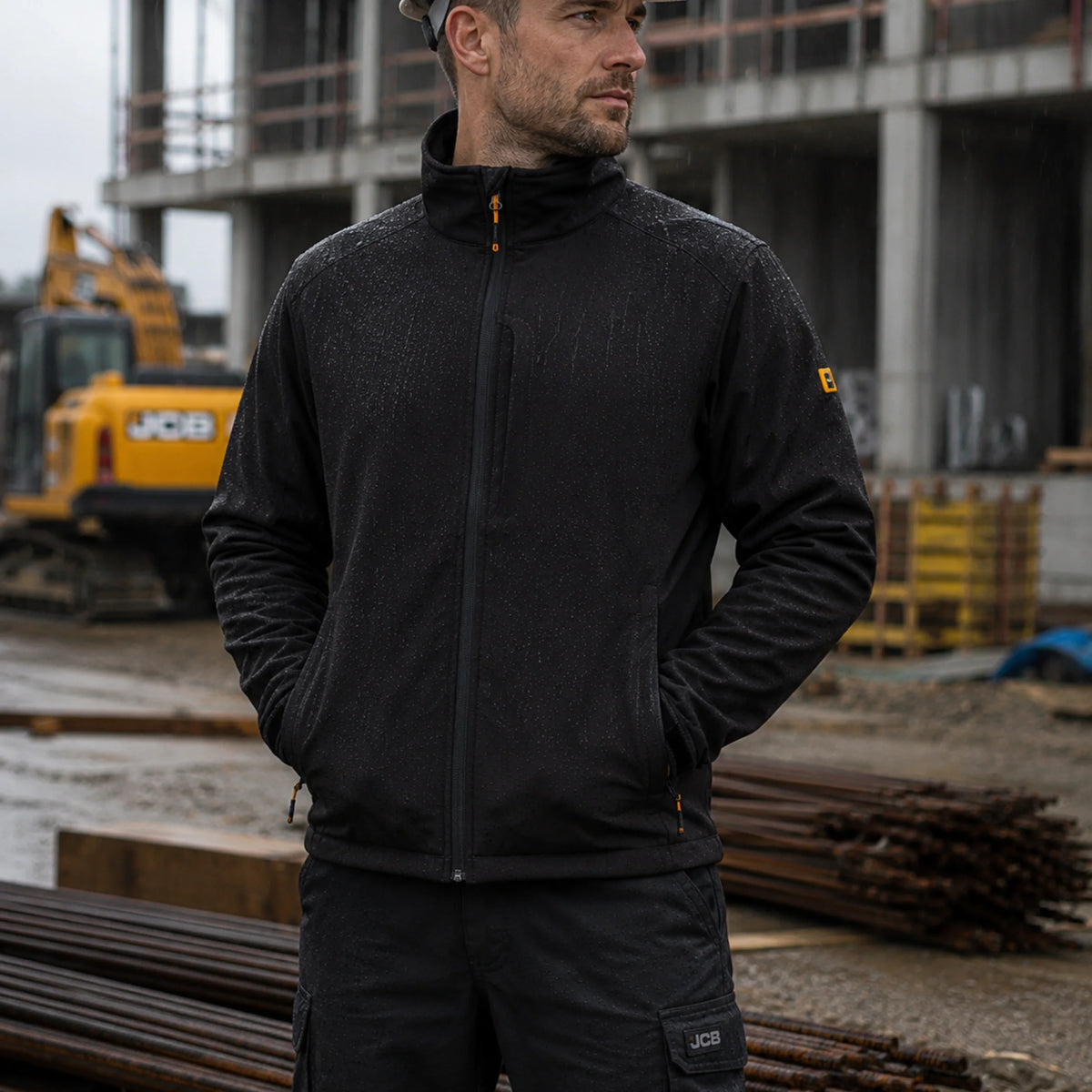 Mężczyzna w czarnej kurtce roboczej softshell JCB na placu budowy z koparka w tle, ujęcie z boku