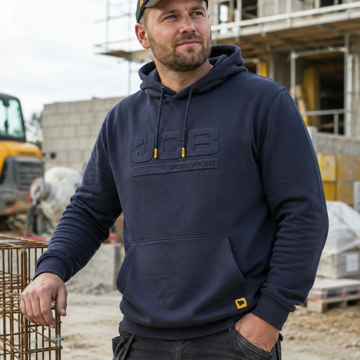 Mężczyzna w granatowej bluzie JCB z kapturem na placu budowy, ujęcie z przodu